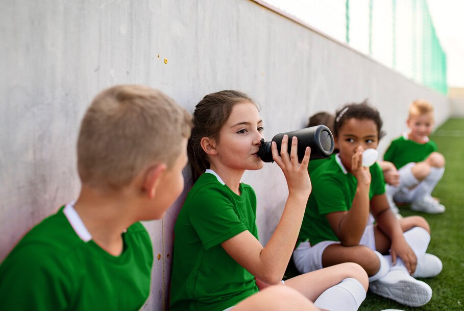 A pee wee sports team sit beside a wall drinking water.